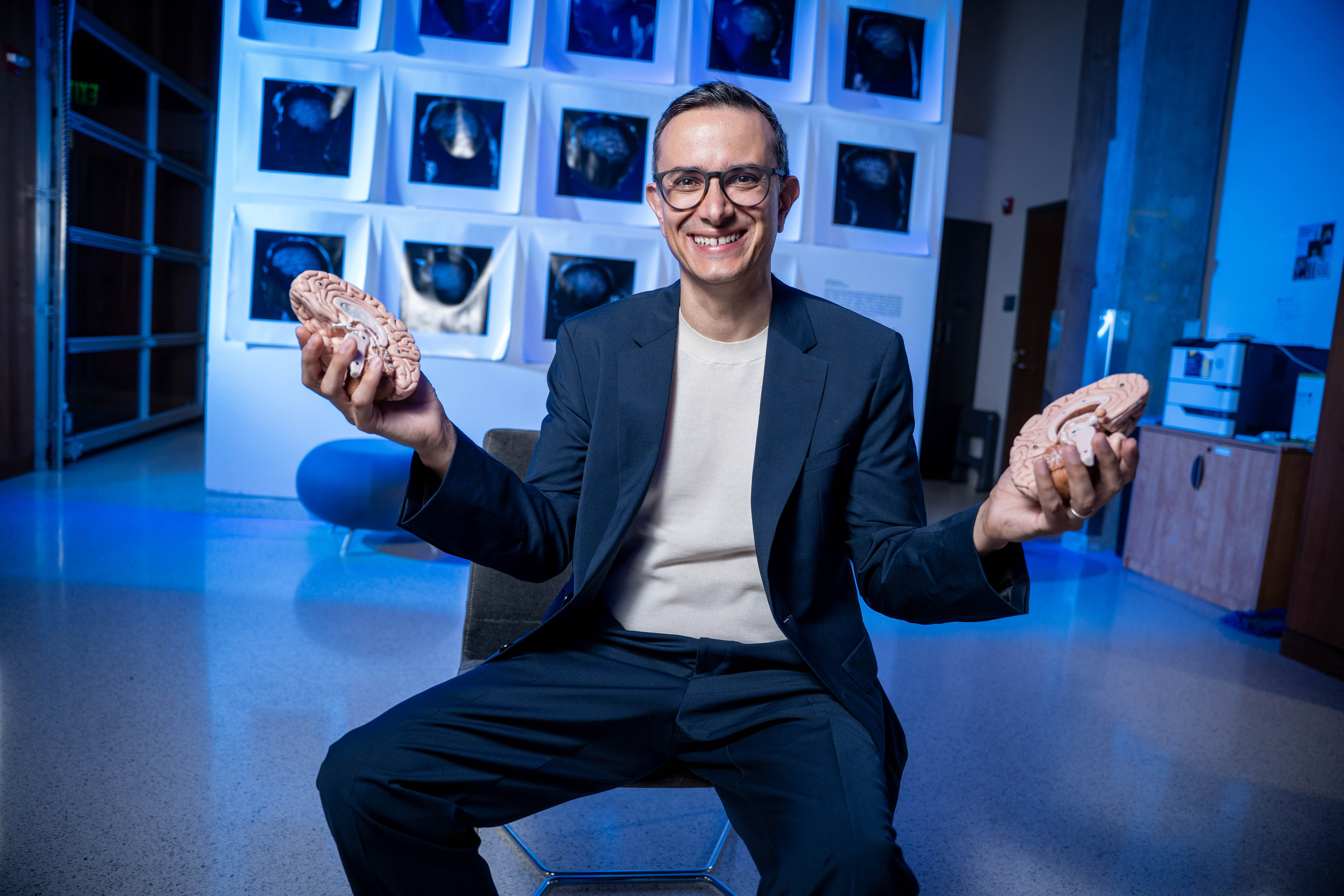 Jorge Morales seated, smiling, holding the two halves of a model brain, in front of a wall of brain scans